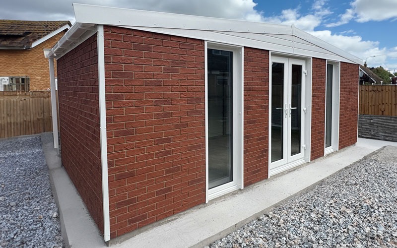 Red brick extension with white-framed glass doors along a concrete patio, surrounded by a gravel yard and wooden fence.