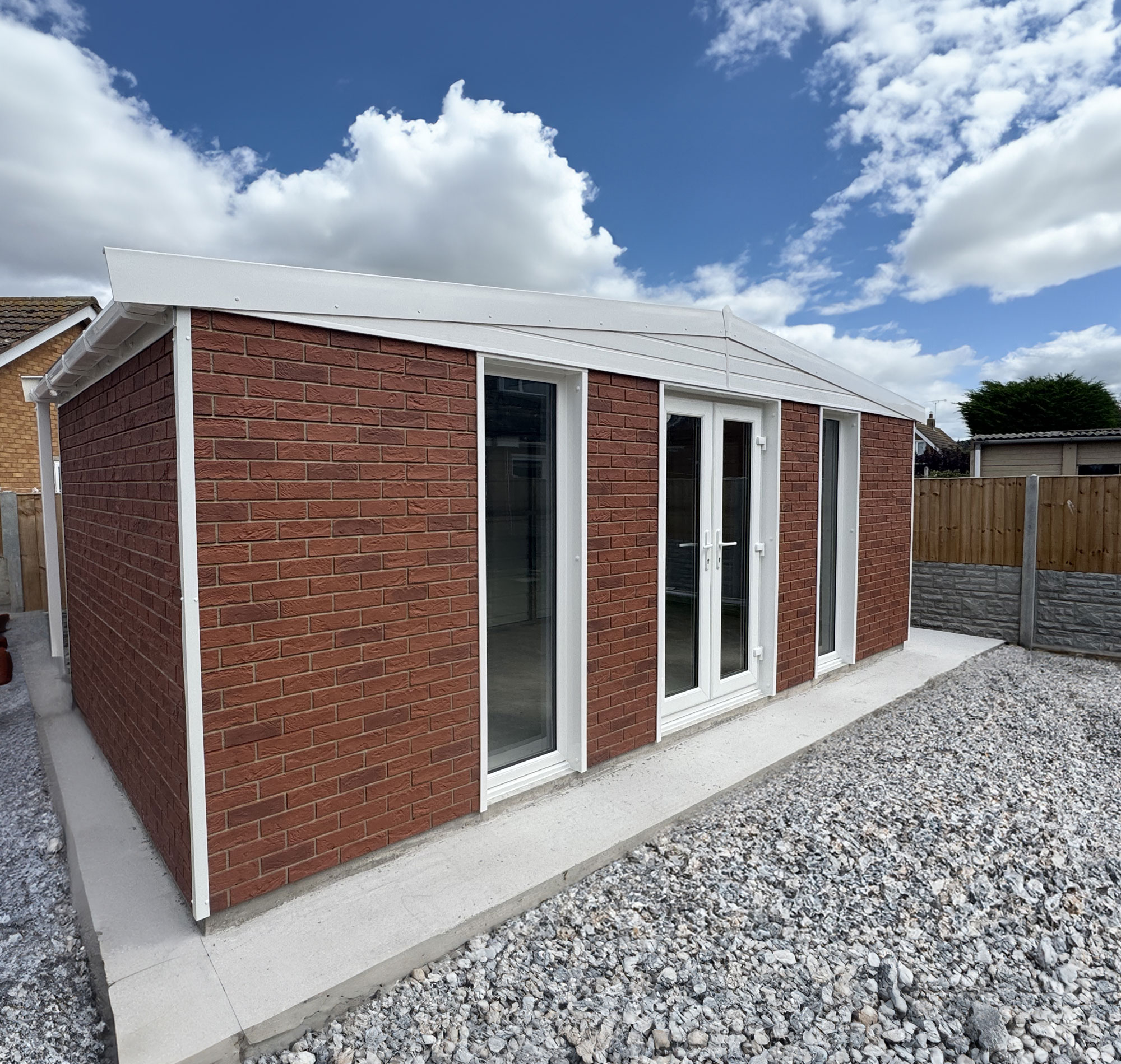 GARDEN-BRICK-MAIN - All Seasons Steel Buildings New brick extension with white-framed glass doors, set on a concrete foundation, surrounded by gravel yard under a blue sky with clouds.
