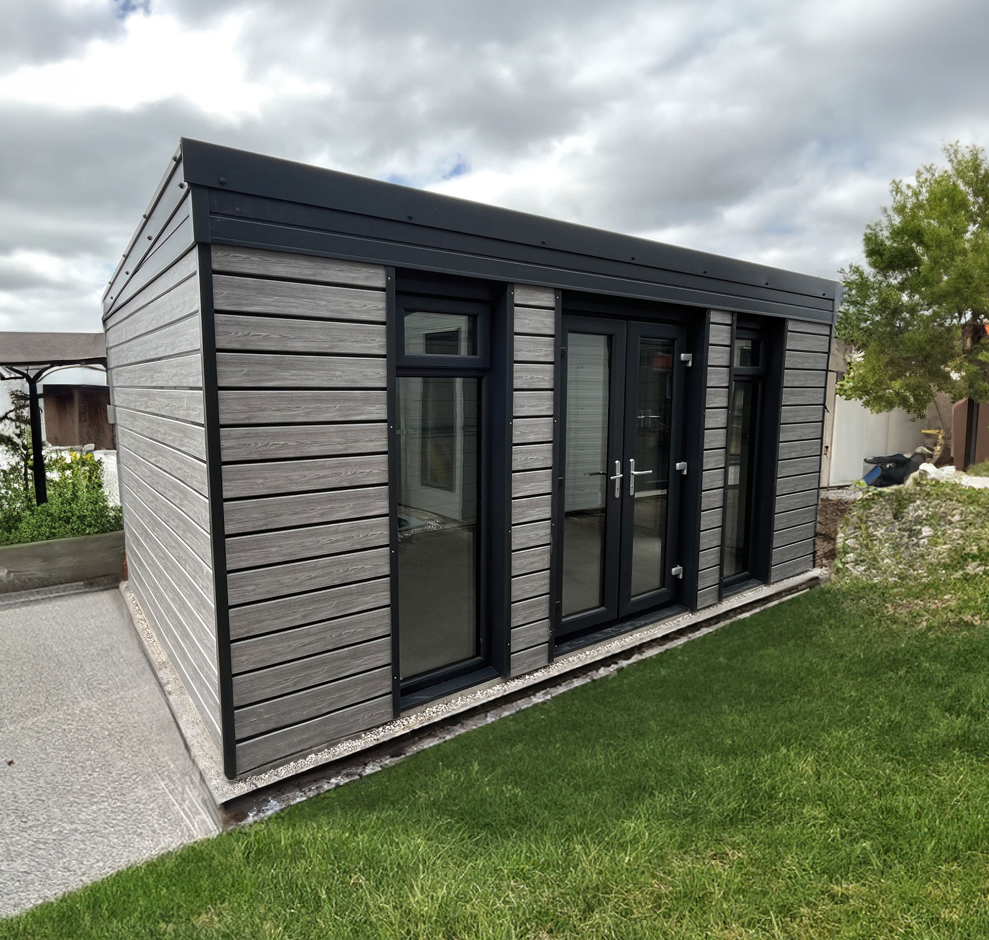 Modern gray wooden shed with large glass doors on a grassy lawn under a cloudy sky.