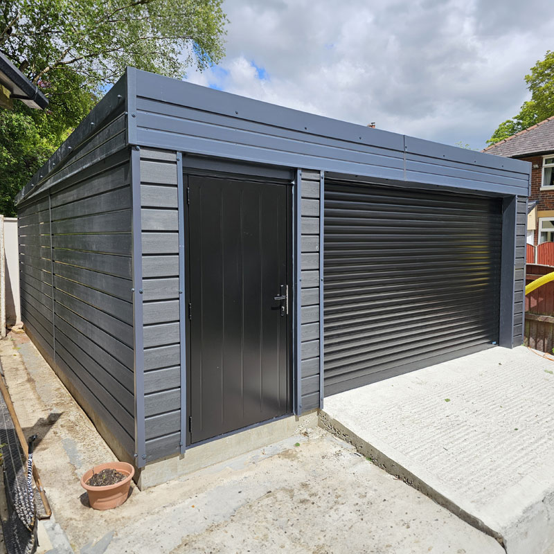 A dark gray metal shed with a single black personnel door on the left and a large black roller shutter door on the right, set on a concrete path outside.