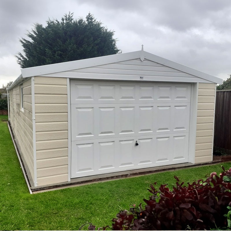 beige detached garage with a white sectional garage door, peaked roof, and small handle, set in a grassy yard on a cloudy day