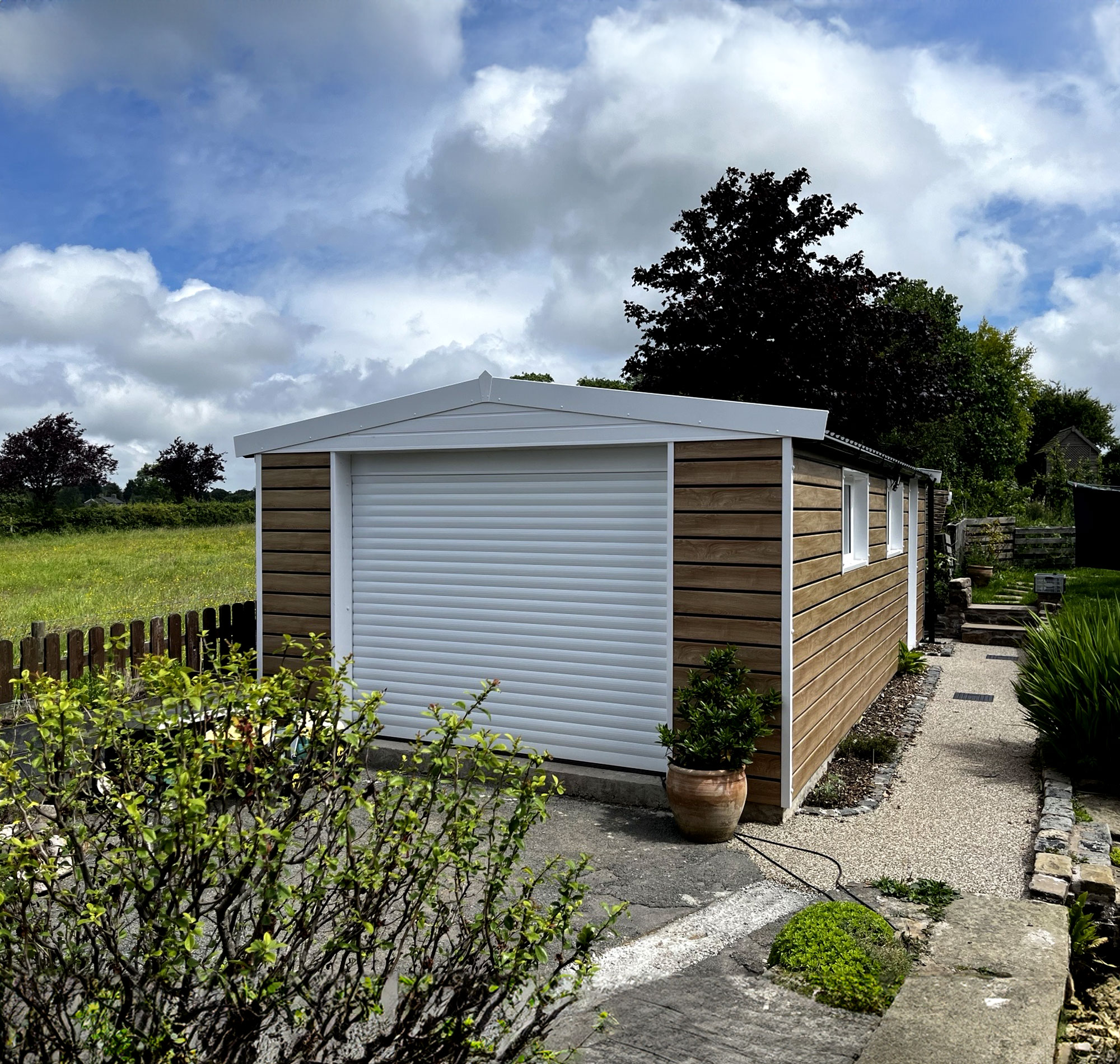 Modern wooden shed with a white roller door, set in a garden with hedges and a cloudy sky.