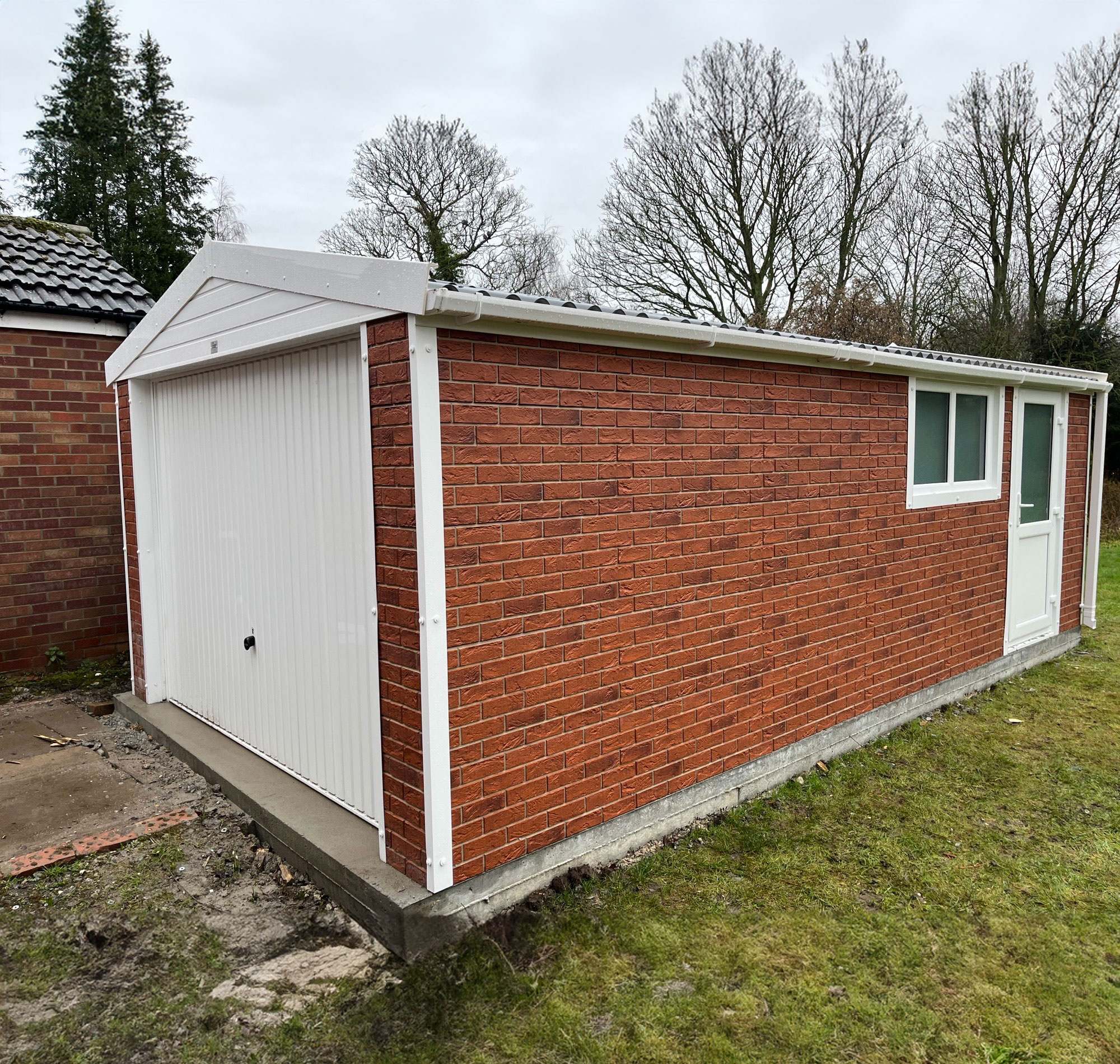 Brick outbuilding with a white, paneled garage door and a small white-framed door/window, set on a concrete base, under cloudy skies.