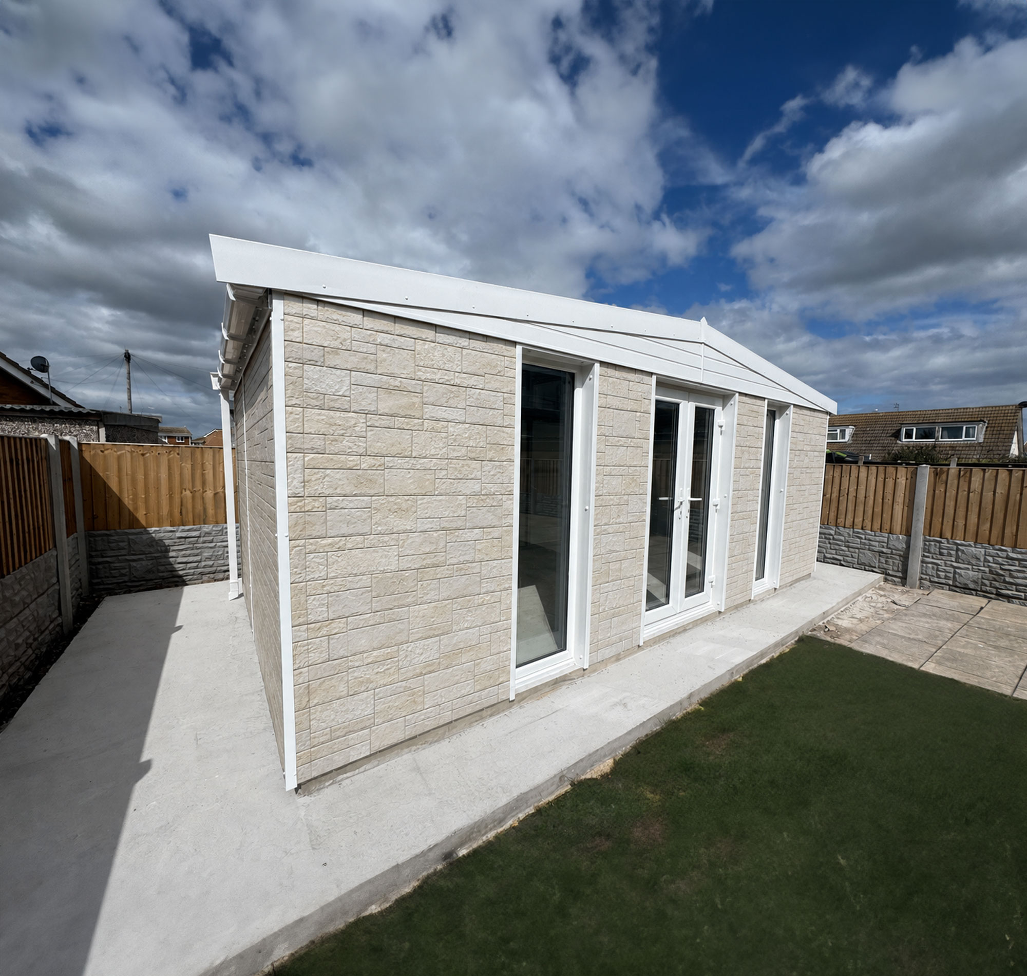 Corner view of a modern beige brick house with white-framed glass doors, a fenced yard, and a cloudy blue sky above.