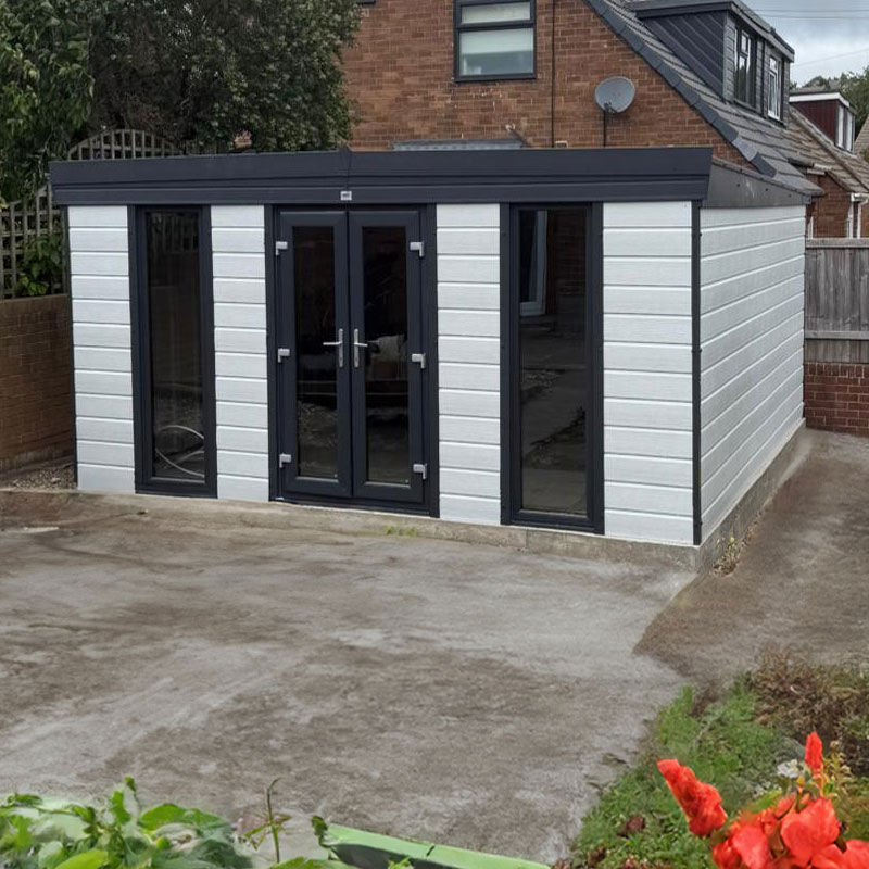 Modern gray garden room with double glass doors and tall side window, on a concrete patio beside a brick house, with red flowers in the foreground.