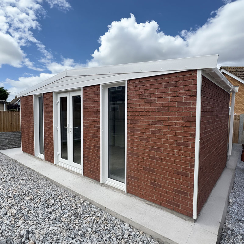 Brick garden outbuilding with white-framed doors and tall window, on a gravel yard under a cloudy blue sky.