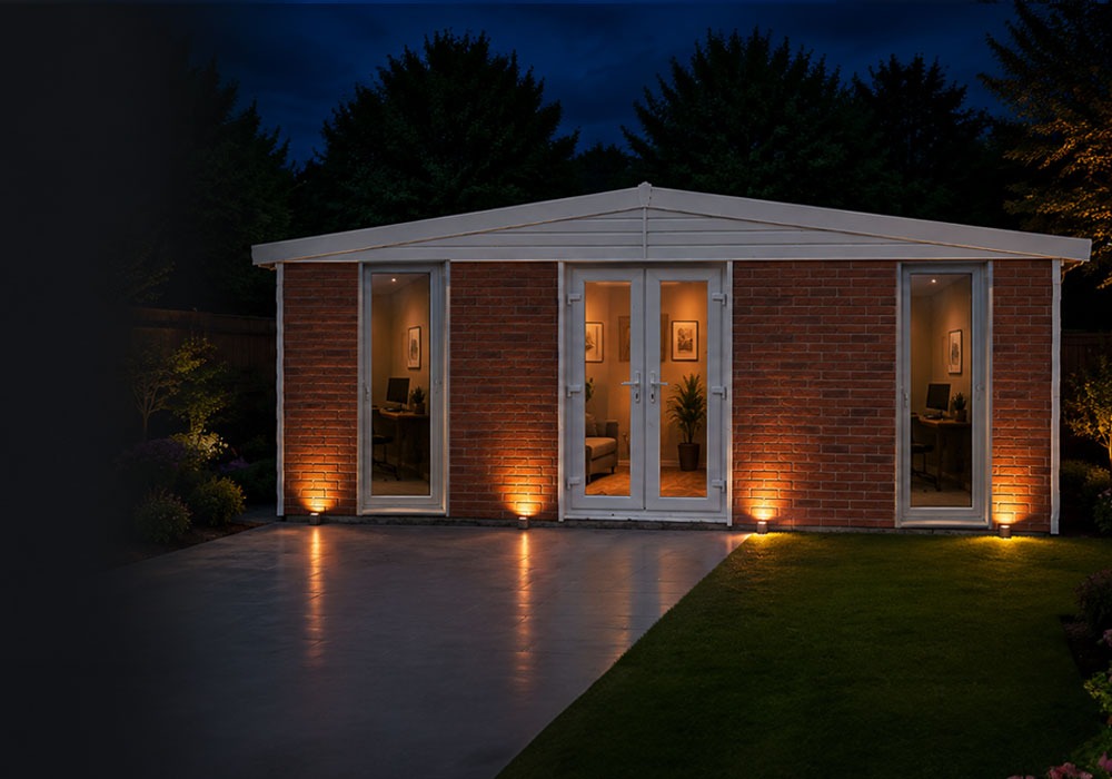 Brick garden studio at night with glass doors, warm interior lighting, and spotlights along the façade.